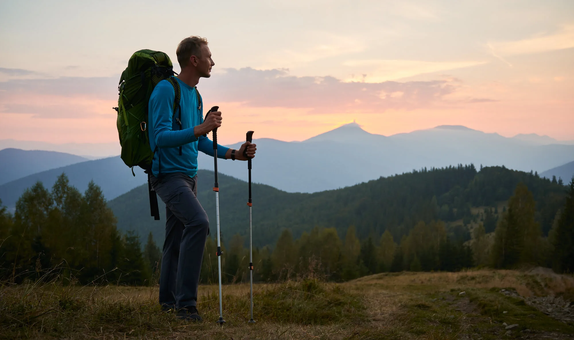Mężczyzna z plecakiem i kijikami trekkingowymi na górskim szlaku o zachodzie słońca – przygotowanie do trekkingu w Toskanii i Słowenii.